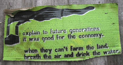 Corrugated cardboard sign, spraypainted green with black smoke-belching factory and words Explain to future generations it was good for the economy. When they can't farm the land, breathe the air and drink the water.