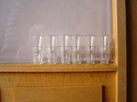 Twelve miniature drinking glasses stacked on the window ledge of a wooden dolls' house shopfront component.