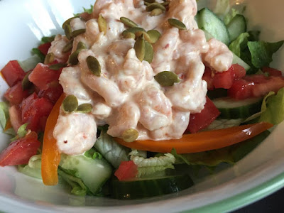 A fresh green salad with visible cucumbers, tomatoes, and orange bell peppers, topped with spicy shrimp salad and roasted pumpkin seeds. 
