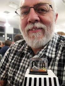 A man behind a  small display box containing the letters 'AMEA', and a tiny cottage and windmill, displayed on an upturned popcorn container.