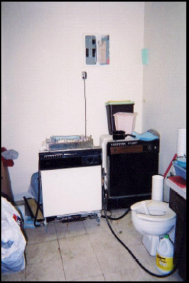 A cluttered room with a portable dishwasher, toilet, trash bags, and unidentified objects on the vanity