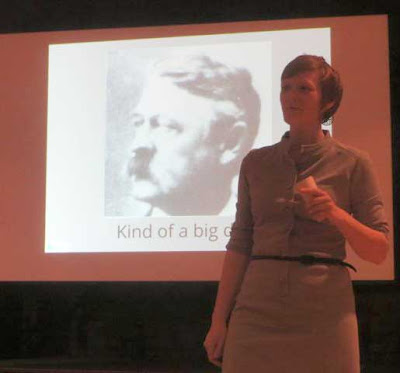Maggie Koerth-Baker standing in front of a projected image of a 19th century old white man with a big mustache