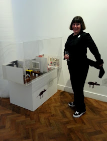 Woman in a gallery, holding  a glass of wine next to an exhibit and showing the tails on her coat.