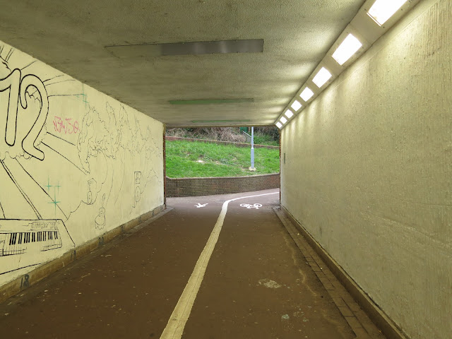 In the underpass. Murals on leaf, lights on right, grass glimpsed through opening beyond.