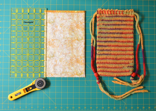 Almost the same as the previous photograph, the lining is laid out on the grid next to the bag.  A quilter's rule is on the left hand edge of the lining to trim the seam allowance. The rotary cutter is pointing to that edge.