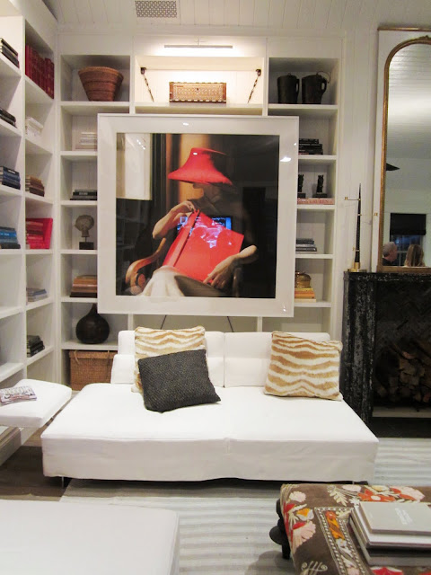 Close up of a piece of art in the Windsor house's family room. It's of a woman in a wood chair holding a red book and wearing a red hat. The piece is in front of a the built in bookshelves and above the what Barcelona club chairs