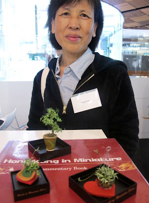 Woman standing in front of a selection of dolls' house miniature plants, arranged on the guest book for the exhibition Hong Kong in Miniature.
