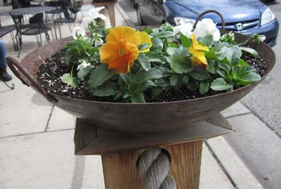 Rusted steel wok full of orange and white pansies, atop a wooden pillar