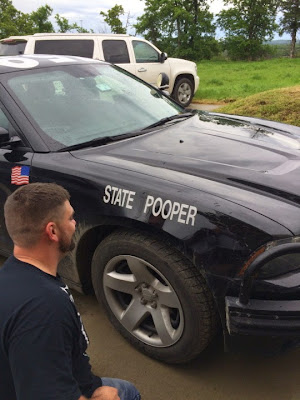 Image of a man kneeling next to a cop car with tape over the T and the leg of the R in "STATE TROOPER" so that it says "STATE POOPER"