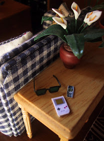 Modern dolls' house miniature sofa table and sofa. On the table is a pot of lilies, a pair of sunglasses, a Gameboy and a Nokia mobile phone.