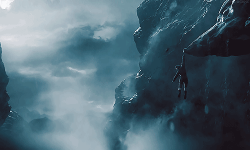 No gif: Lara Croft pendurada com uma mão só na ponta de um penhasco em um ambiente todo cinzento com nuvens no fundo brilhando com relâmpagos e névoa ao redor
