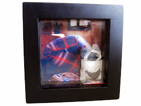 A box picture frame containing a photo of a modern miniature scene, and a miniature knitting bag and book included in front of the photo.