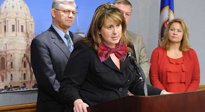 Photo of Amy Koch speaking at a podium, wearing a dark suit with a Photoshopped bright pink plaid bow tie