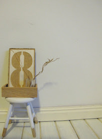 Modern dolls' house miniature stool with raw wood legs and white-dipped top part., displaying a letter eight in a wooden box, along with a glass with twigs in it.