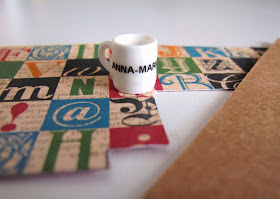 A piece of scrapbooking paper printed with squares containing typography. On it is a dolls' house miniature mug, and next to it is a piece of brown cardboard.