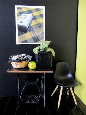 Modern dolls' house miniature scene of a vintage sewing table and Eames chair in front of a black wall displaying a poster with vintage pins on a checked fabric.