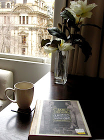 Table with a vase of magnolias, a mug and a book on it. The window behind overlooks an old Melbourne building on Swanston Street..