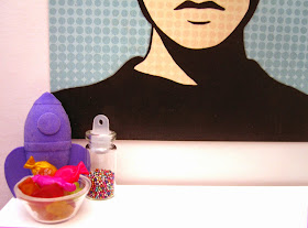 Modern dolls house miniature scene of a purple rocket, bowl of colourful sweets and jar of tiny beads in front of a colourful poster.