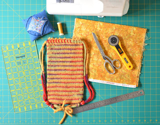 Top view of materials and tools: sewing machine, quilters' rule, pincushion with pins, reel of sewing thread, swatch of cotton fabric, scissors, rotary cutter, steel rule and WIP Project Bag on a cutting mat.