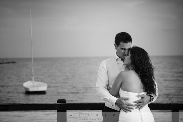 novios abrazados apoyados en puente de madera junto un barco sobre el mar