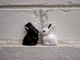Two foil-covered chocolate bunnies sitting on a white wall. One has been painted black, the other, white.
