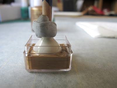 White dolls' house miniature vase, with the end of a paint brush blue tacked into it, dipped into a small plastic container half-filled with gold nail varnish.