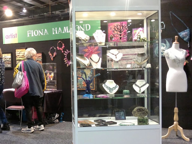The right hand side of Fiona Hammond's exhibition contains a glass display cabinet containing jewellery and sculptural works. A dressmaking mannequin on the far right displays wearable art pieces with fashion accessories like bags on the wall behind. On the left hand side of the photograph a visitor is viewing the necklaces on the back wall underneath the green sign and inspecting the contents of a smaller display cabinet towards the back of the exhibition space.