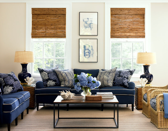 Living room with two navy sofas with white piping and blue and white floral and chevron printed accent pillows,two wicker armchairs with blue cushions, a wood coffe table with metal legs, matching wicker side tables with black lamps and encasement windows