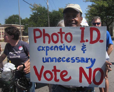 Man holding hand-painted sign reading Voter ID is expensive unnecessary VOTE  NO