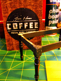 Miniature dining table leg assembly, painted black, sitting on a cutting mat with a spray can of chalkboard paint and a wooden coffee storage box.
