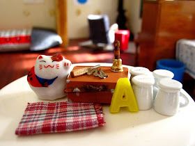Selection of modern miniature homeware items displayed on a tabletop, including a tea towel, set of mugs, old school case, bell, letter A and cat ornament.