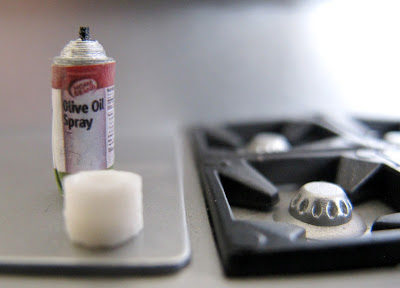 Modern dolls' house miniature bottle of olive oil spray on a bench next to a stovetop.