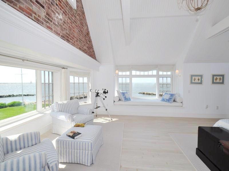 Bedroom with light wood floor, exposed brick walls, striped upholstered armchair and ottoman, a black chest and a large picture window with a view of the Long Island Song. There is a smaller window with a window seat and encasement windows to the left of the picture window.