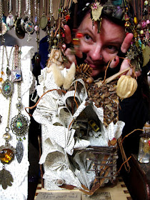 A woman peeks through a display of necklaces on a market stall above a paper house.
