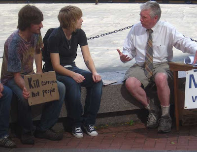 Two young guys talking to a guy in a tie, one young guy with small sign that says Kill corruption not people