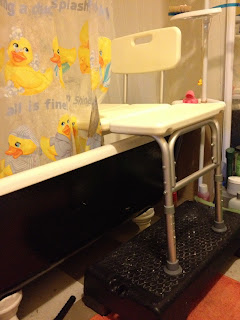 Photo shows well-lit bathroom with clawfoot tub, shower chair in the tub, sitting on a black aerobics step. Also visible is the rubber ducky shower curtain and an orange bathmat under the step