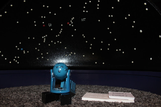 Ready for my close-up! The model is now out in the Planetarium's lower level in the lobby! The black dome with all the stars of white rivets sits upon the purple wall and grey carpet. In the middle is the Zeiss model. The teal oval StarBall on stilts sits on the two smaller black wedges and the teal eight planet projector vertical boxes sit on the long black wedge. Next to it on the carpet sits a small white exhibit text panel of about three inches by one-and-a-half inches. It has Braille and large print marking the StarBall as exhibit one and the Planet Projectors as exhibit two to correspond to the other exhibit text.