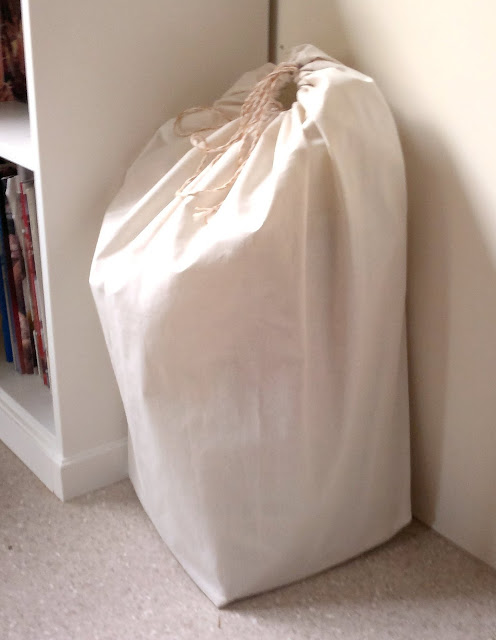 A calico drawstring bag in the corner next to the bookcase. The neutral colour blends in to the decor and hides a mini bale of oaten hay.  There is a circular opening at the top. It is drawn closed by a home-made twisted cord.