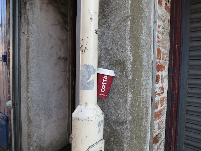 Cost Coffee cup stuck between a lampost and a concrete wall.