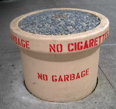 Large concrete cylinder, about three feet tall, full of round rocks, stencil letters on the side saying No Cigarettes No Garbage