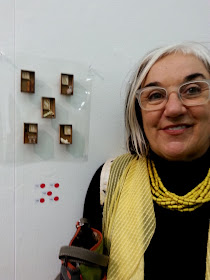 A woman stands in front of five miniature brooches on display in a gallery, with five red dots underneath.