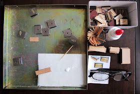 A magnetic gluing jig, box of wooden alphabet  tiles, glue, toothpicks, kit pieces, instructions and reading glases laid out on a dining table.