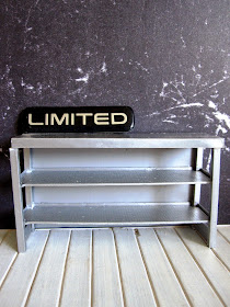 Modern doll's house miniature industrial grey storage bench in front of a wall covered with black marble-patterned paper. On the bench is a sign saying 'Limited'.