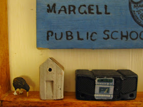 A small clay house displayed in a dolls' house between a kiwi figure and a portable tape deck and CD player.