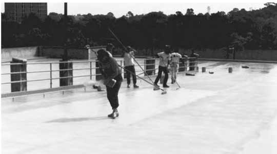 Deck-coating application. Note the pushing of squeegees in the background, back-rolling of coating and spreading of the aggregate by hand in the foreground. All crew members are wearing golf shoes. Deck-coating application. Note the pushing of squeegees in the background, back-rolling of coating and spreading of the aggregate by hand in the foreground. All crew members are wearing golf shoes.