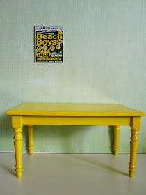 Bright 1960s Beach Boys poster on a green wall with a modern dolls' house miniature yellow table underneath,