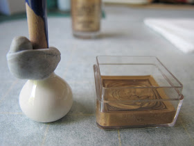 White dolls' house miniature vase, with the end of a paint brush blue tacked into it, on a piece of baking paper next to a small plastic container half-filled with gold nail varnish.