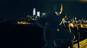 Photo of Daredevil character, in black suit with mask over most of face, in front of nighttime NYC skyline