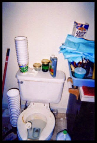 Dirty toilet with stacks of empty styrofoam cups on and beside it and assorted clutter on all sides