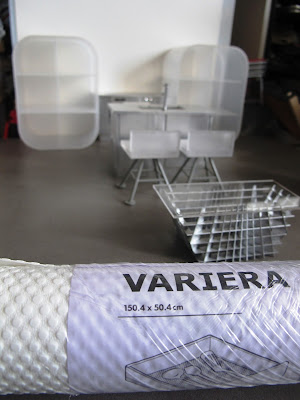 Modern dolls' house miniature white and grey kitchen set up, with concrete floor. In the foreground is a roll of white IKEA VARIERA drawer mat.